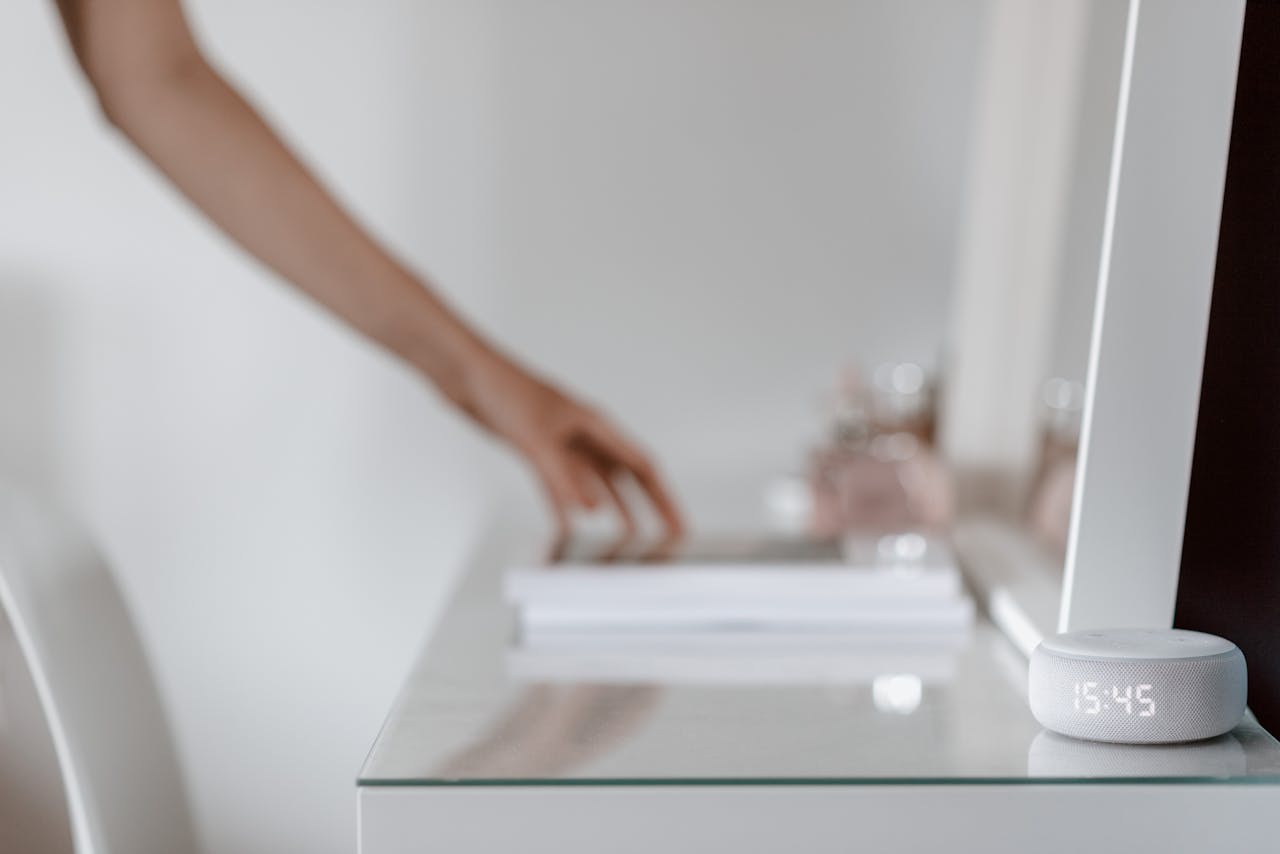 A smart speaker with a digital clock display placed on a minimalist desk setting, hand in background.